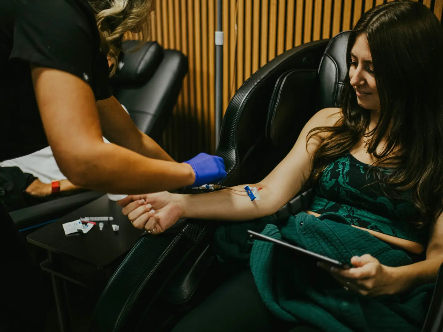 Woman receiving IV drip in her arm from a healthcare professional, holding a tablet